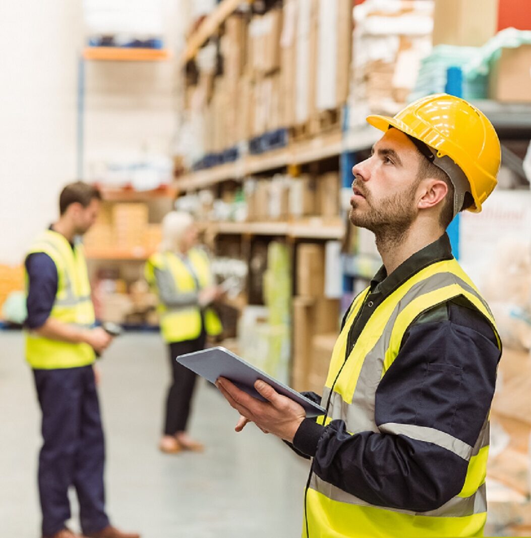 A man in a hard hat and safety vest surveys the warehouse, ensuring safety and compliance in the work area.